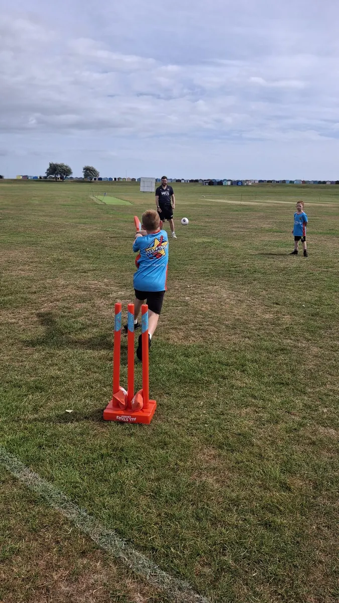 Young All Stars batter in blue mid-swing at orange stumps on a grass field