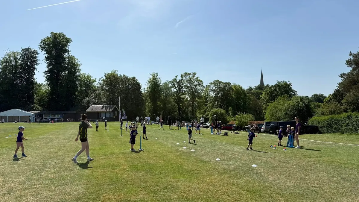 Children in purple kit doing junior cricket drills at a sunny club ground with a pavilion and church spire behind