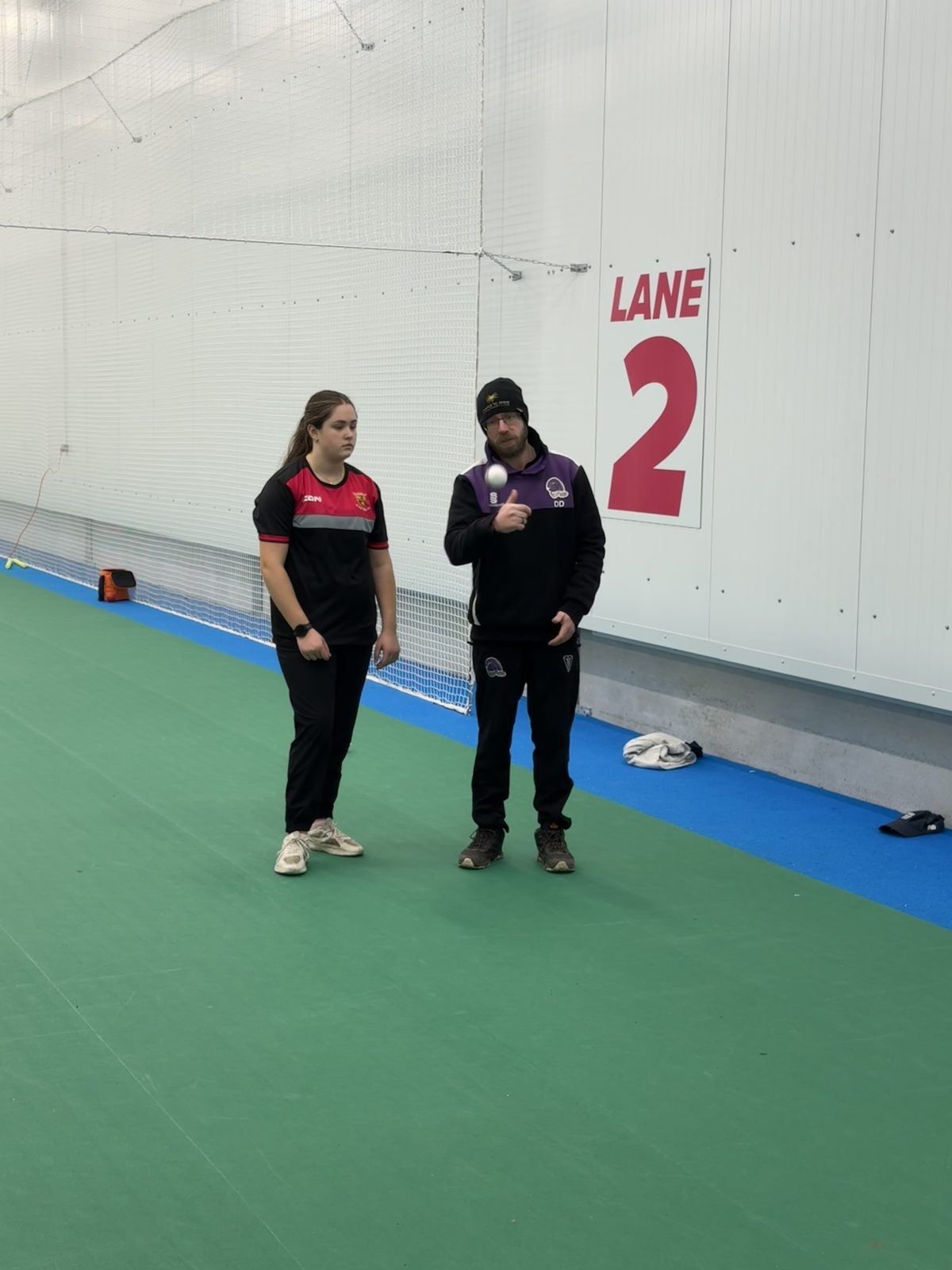 Coach spinning a cricket ball beside a young player under a lane 2 sign in an indoor centre